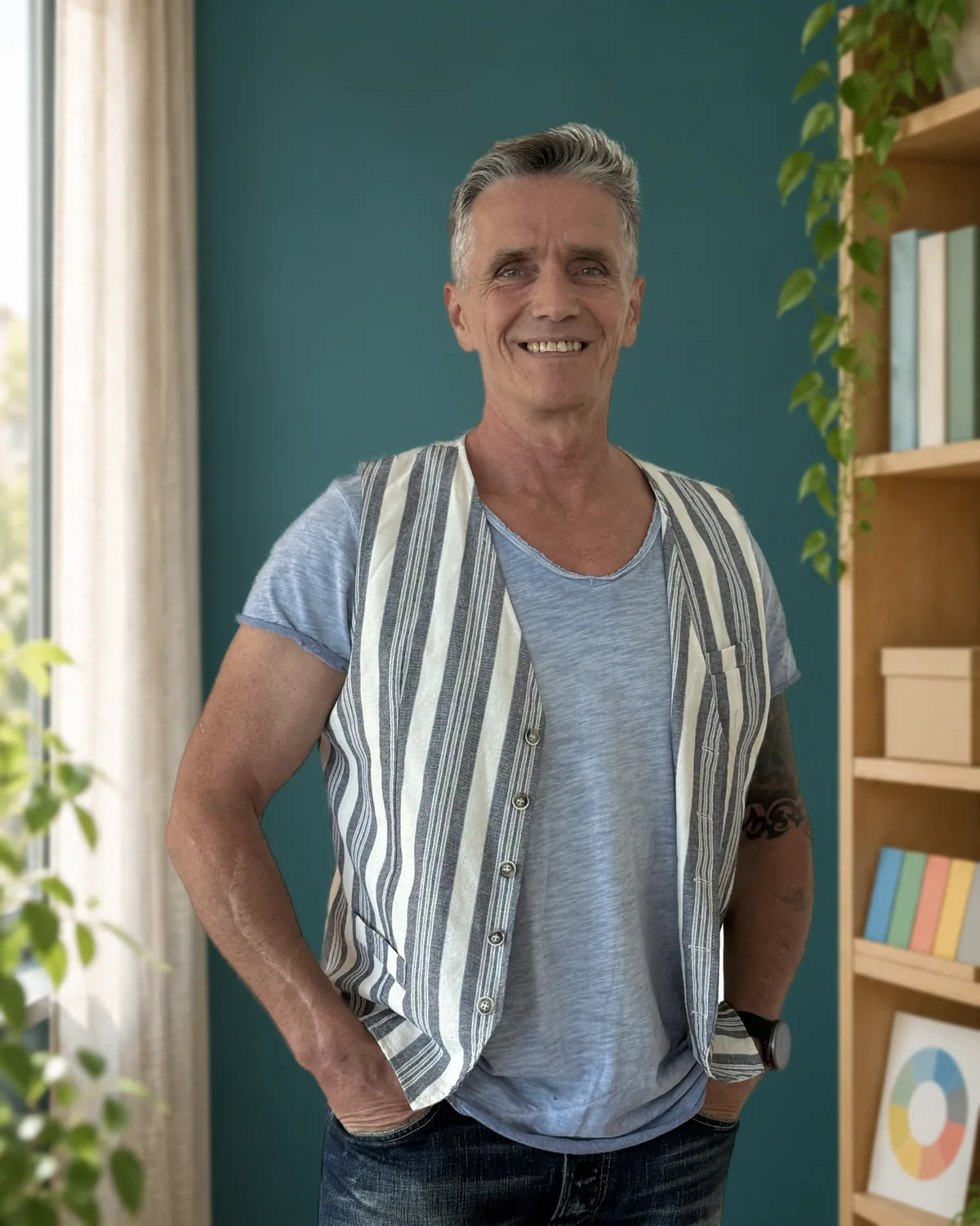 Ron Jansen in gestreifter Weste, eine Hand in der Hosentasche, lächelt vor einer petrolfarbenen Wand; daneben ein Holzregal mit Büchern und Pflanze.