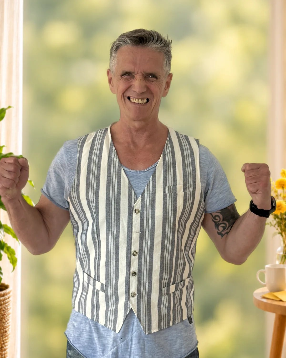 Ron Jansen lacht und ballt beide Fäuste vor Freude; hinter ihm ein heller Raum mit Blick ins Grüne und gelben Blumen.